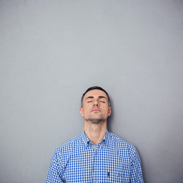 Man standing against a grey wall with his eyes closed; thinking.