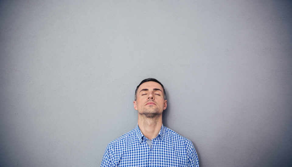 Man standing against a grey wall with his eyes closed; thinking.