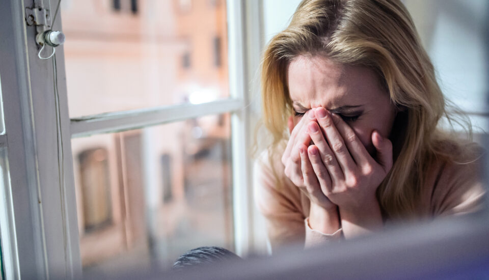 Woman sitting on windowsill, looking out of window, crying.