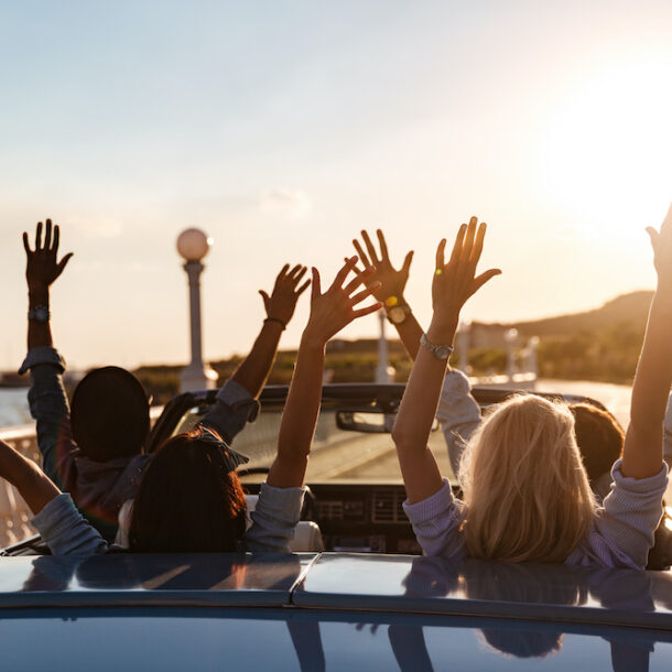 Back view of happy friends driving in a convertible with raised hands.