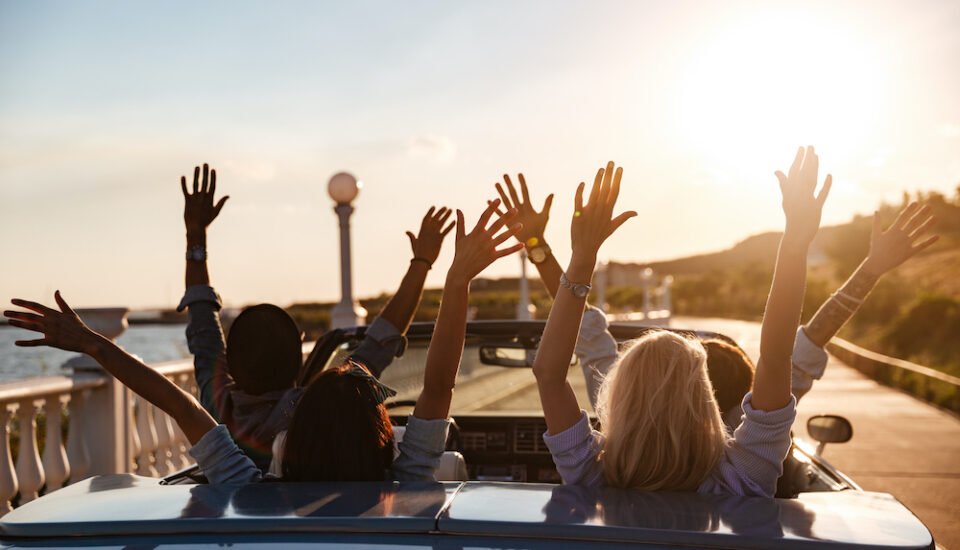 Back view of happy friends driving in a convertible with raised hands.