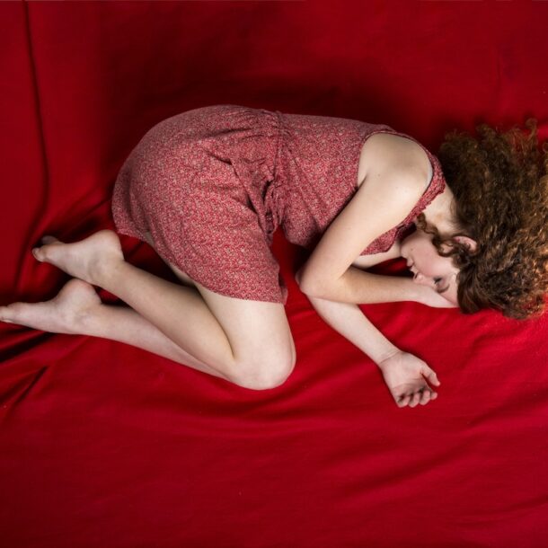 Young, sad woman in fetal position laying on a red background.