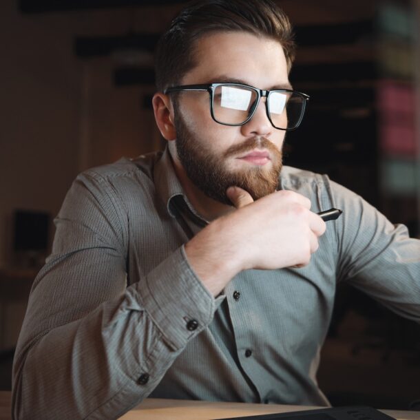 Photo of young professional dressed in shirt and wearing eyeglasses working late at night and looking at computer.