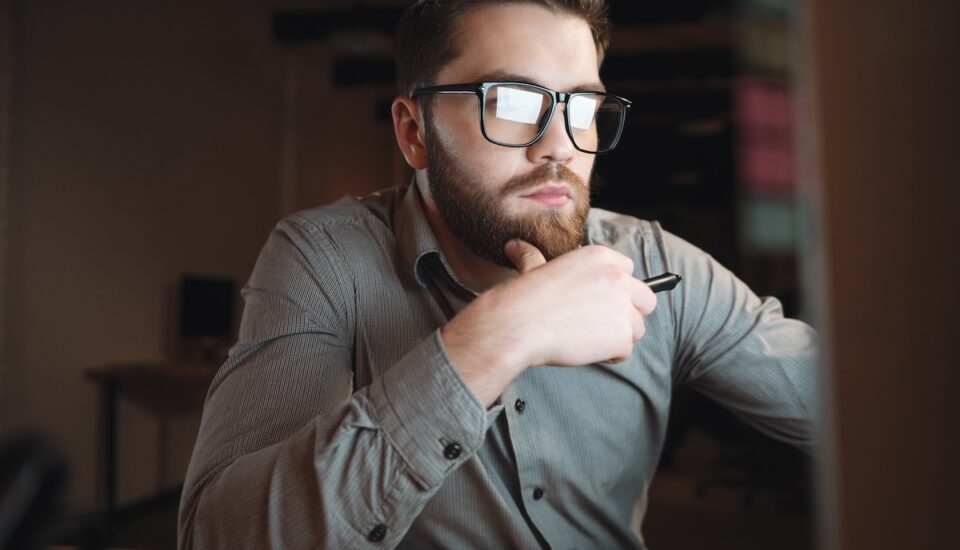 Photo of young professional dressed in shirt and wearing eyeglasses working late at night and looking at computer.